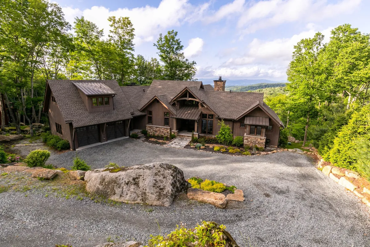 The exterior of Twin Boulder Lodge is showcased, featuring the house set among large boulders, a landscaped garden, and a spacious driveway. The inviting porch hints at opportunities for family gatherings and outdoor fun.