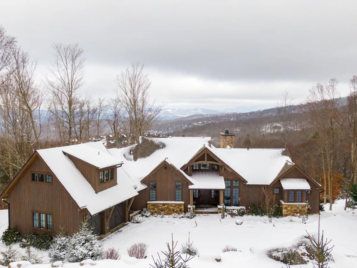 An exterior winter view of Twin Boulder Lodge, showcasing the main entrance, a prominent stone chimney, large windows, and a snow-covered roof. The scene evokes a sense of coziness and warmth, even as snow blankets the landscape, suggesting proximity to winter sports and après-ski relaxation.