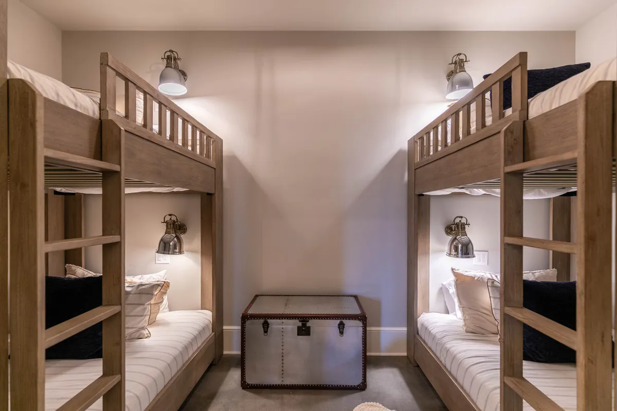 A playful, inviting bunk room featuring sturdy wooden bunk beds, a wooden trunk, and warm wall sconces. The space is designed for children or teens, with cozy beds and thoughtful touches that create a fun, relaxing environment.