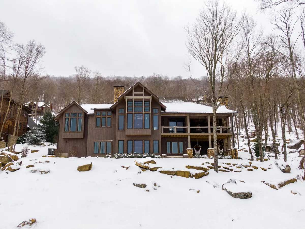 A wintery scene featuring the home’s large windows, a wooden exterior, a snow-covered ground, and nearby trees and rock formations. The inviting porch looks out over the winter landscape, evoking cozy indoor-outdoor transitions.