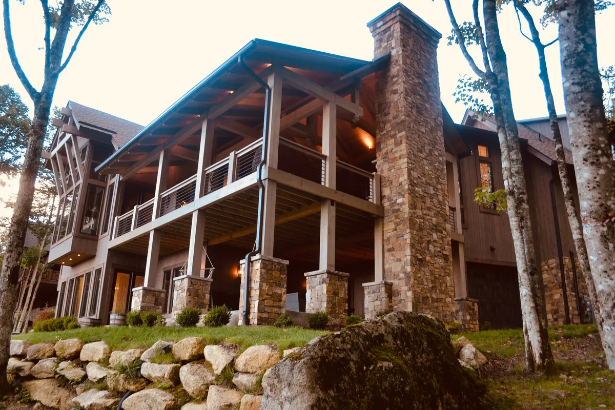 This exterior image captures the large stone pillars and expansive porch of a mountain home, with detailed woodwork and lush surrounding greenery. The scene evokes gatherings on the porch, evening meals, and tranquil moments overlooking nature.