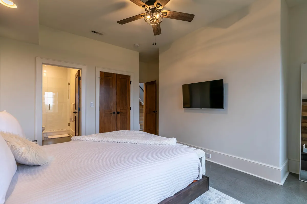 A bright, serene bedroom with a comfortable bed, ceiling fan, television, and a door leading to an adjacent bathroom. Natural light streams through the windows, creating an inviting atmosphere for both rest and entertainment.