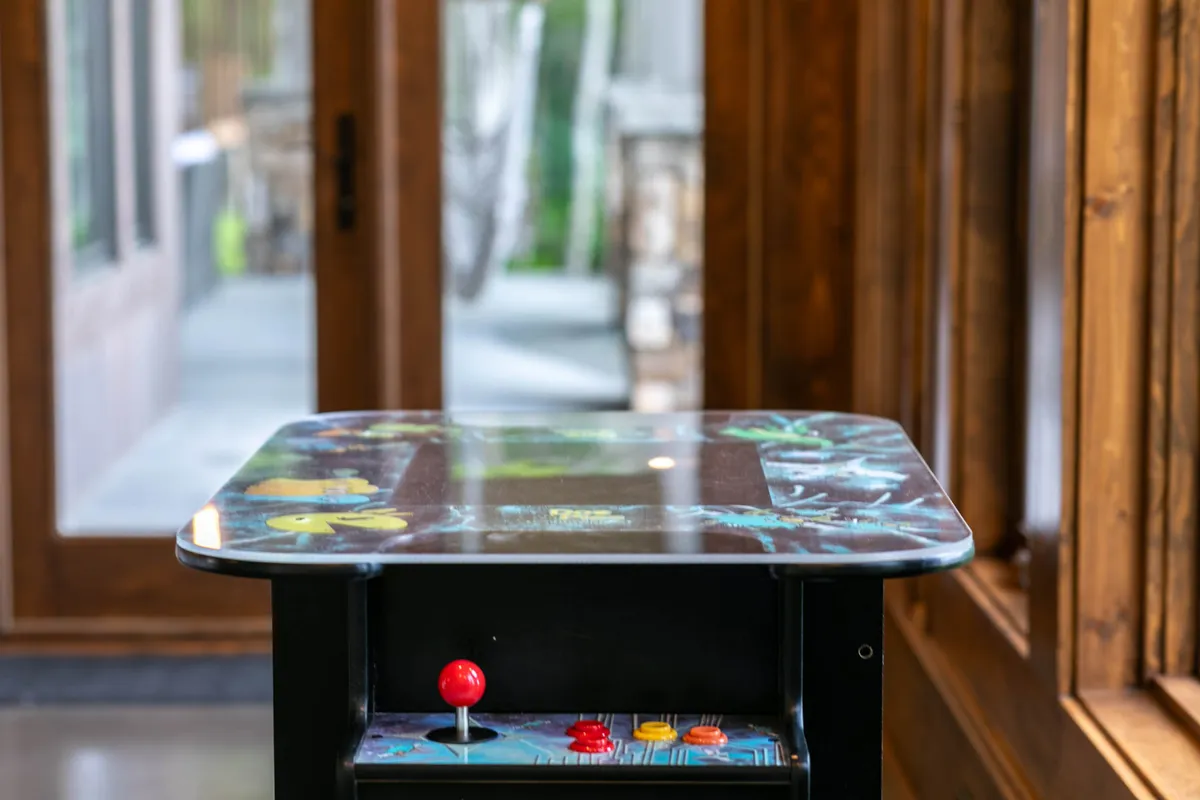 A bright recreation room featuring a retro arcade game table in the foreground, an open doorway, and natural light streaming in from an unseen window. The space exudes a playful, nostalgic energy, inviting guests to enjoy classic video games and friendly competition.