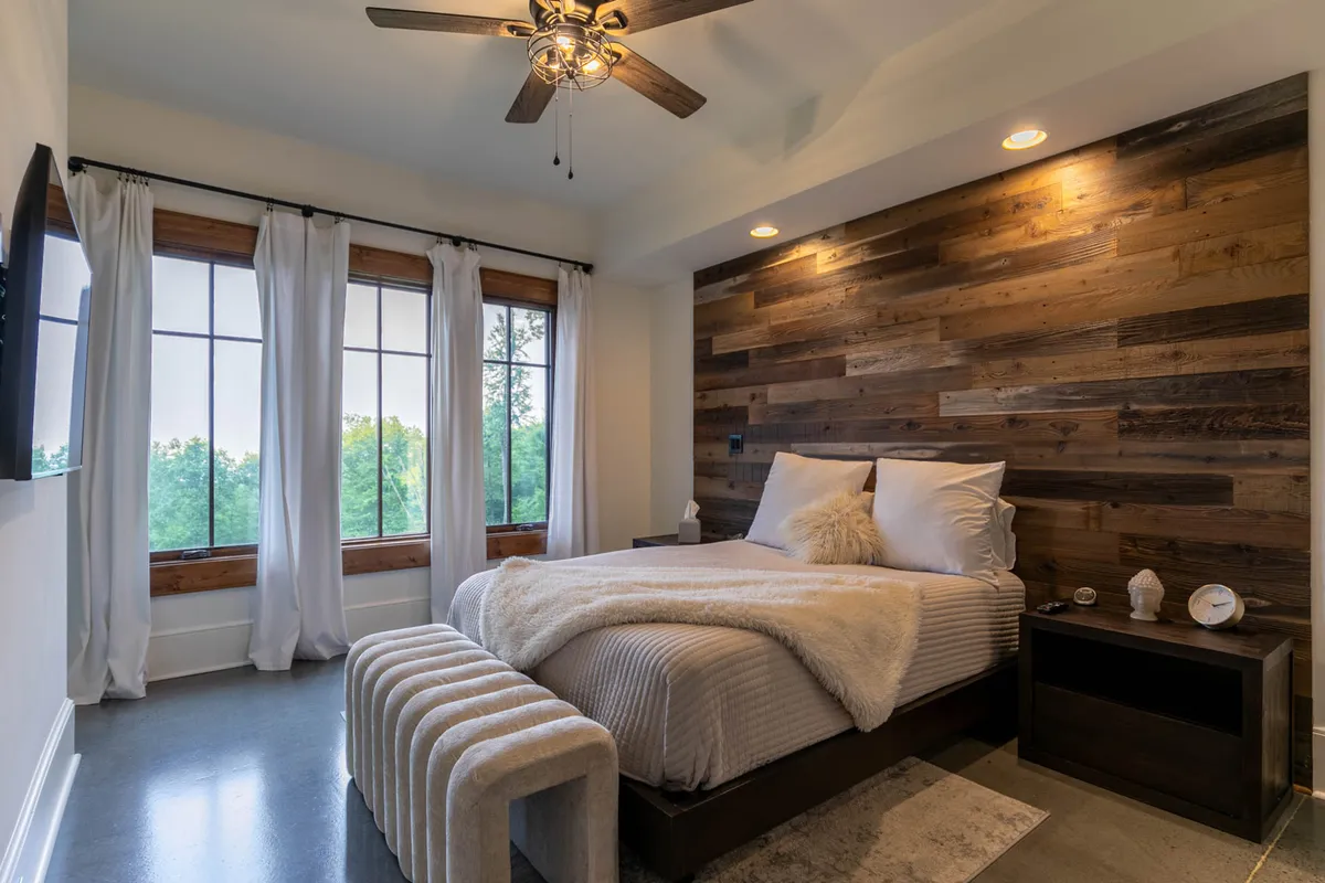 This image features a cozy bedroom with a wood accent wall, large windows letting in natural light, a bed with fluffy pillows, and a ceiling fan. The soft tones and plush bedding evoke comfort and tranquility, making it an ideal representation of a restful retreat.