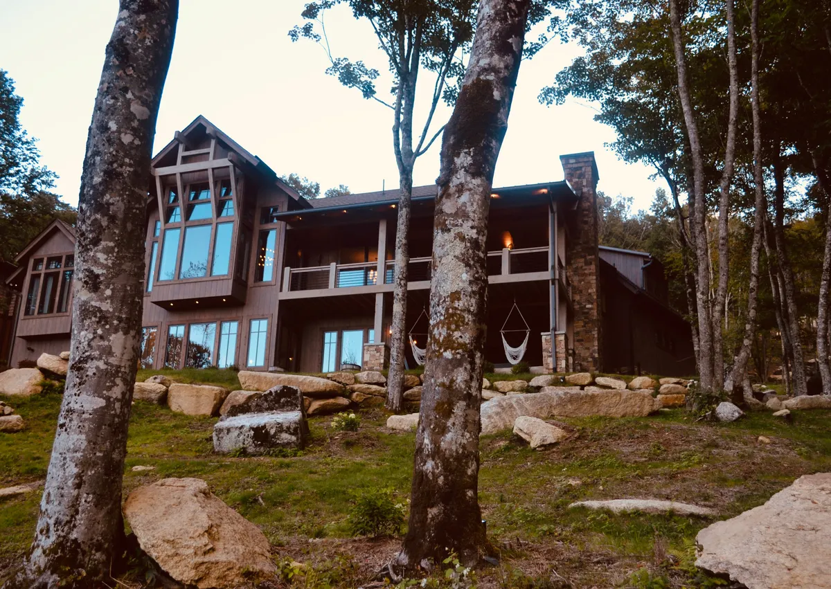 This image captures the large windows of Twin Boulder Lodge, a hammock on the porch, a stone pathway, and natural boulders in the foreground, all framed by tranquil natural surroundings.