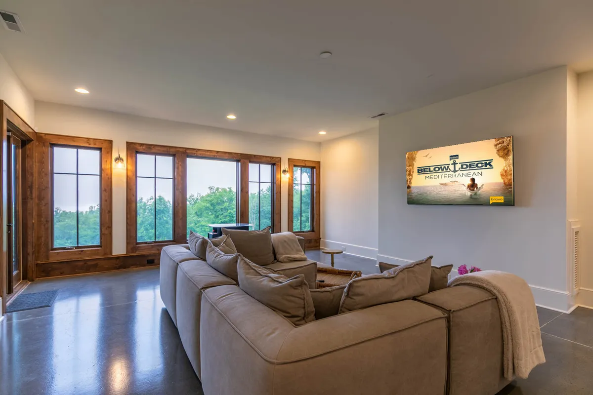 A cozy entertainment space with a large sectional sofa oriented towards a wall-mounted television, which displays a vibrant show. Large windows on one side provide scenic views and natural light, creating an inviting setting for movie nights or casual relaxation.
