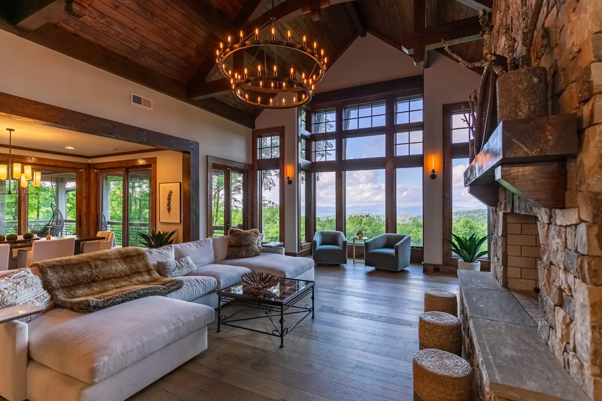 A spacious great room with a comfortable sectional sofa faces a dramatic stone fireplace and towering windows offering breathtaking mountain views. A rustic chandelier and exposed beams enhance the lodge's warm, inviting ambiance, while the natural light fills the space, highlighting both the cozy seating area and the scenic landscape beyond.
