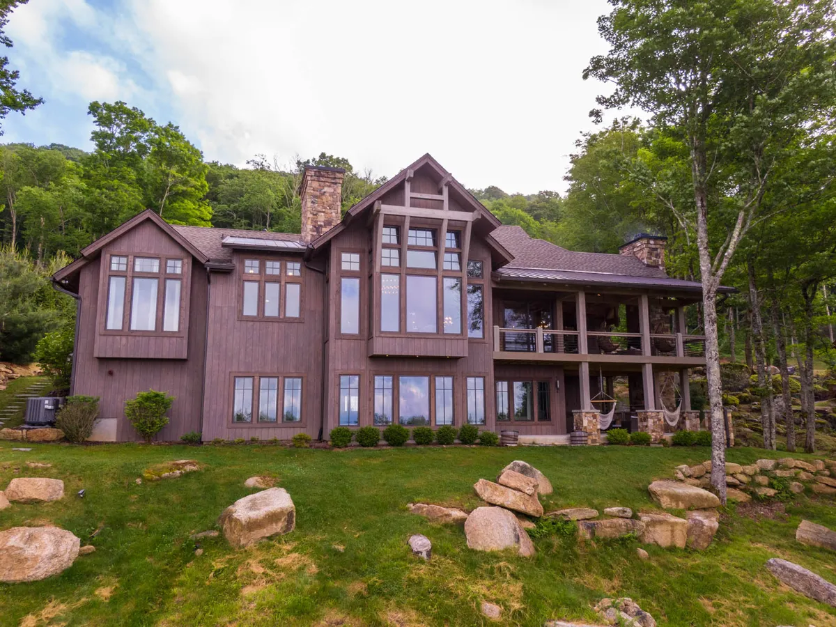 The hero image showcases the exterior of Twin Boulder Lodge: a stunning mountain home with expansive windows, a grand stone chimney, a spacious wooden deck, and a beautifully landscaped garden nestled against breathtaking long-range mountain views. The setting suggests peaceful outdoor living, with areas for gathering around the fire, relaxing on the deck, and soaking in the serene natural surroundings.
