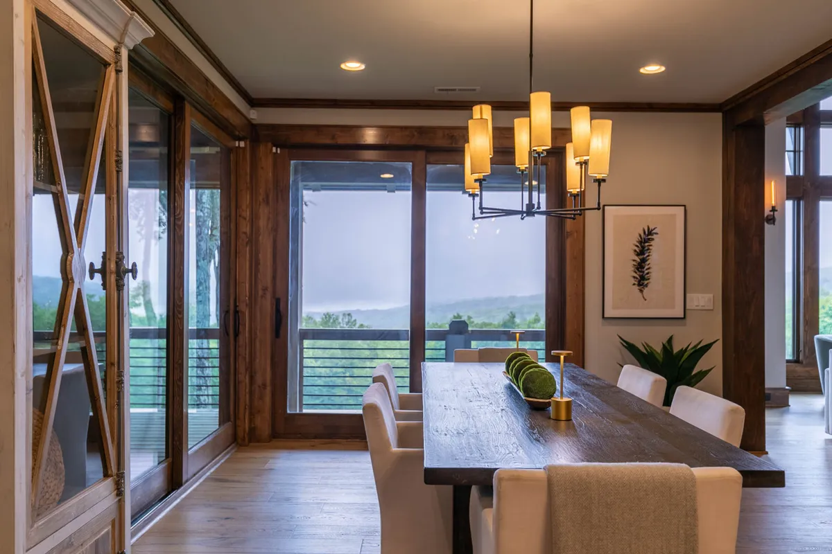 A large wooden dining table sits beneath a warm chandelier, surrounded by windows that frame the picturesque outdoor scenery. The setup exudes a welcoming mood perfect for family dinners, celebrations, or casual gatherings, with natural light enhancing the ambiance.