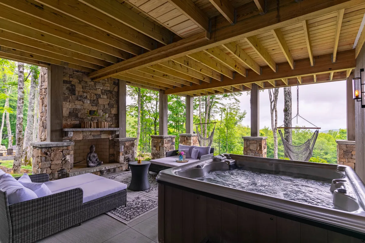 This exterior image prominently features a spacious deck area with a bubbling hot tub, inviting outdoor seating, a stone fireplace, and hammocks surrounded by lush greenery. The setting suggests relaxation in a private, elevated outdoor living space that blends seamlessly with the natural landscape.