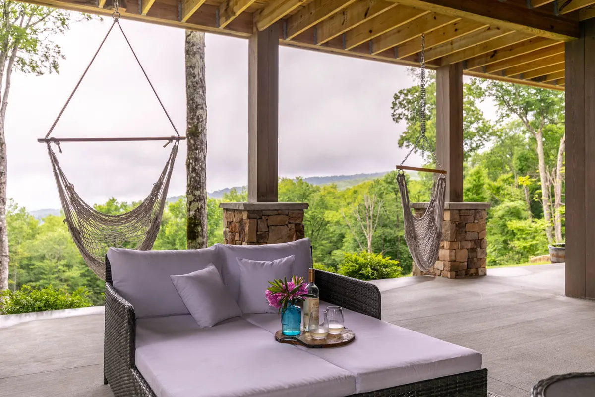 A serene exterior setting with a hammock, sofa, and a table topped with flowers, all arranged to enjoy the natural surroundings. The mood is calm and inviting, suggesting peaceful relaxation or social moments outdoors.